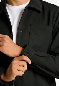 Camisa negra de botones con cuello, que cuenta con un bolsillo en el pecho y puños abotonados. La tela parece texturizada y suave.