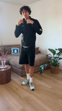 Νεαρός ρυθμίζει μαύρο μπουφάν, φοράει μαύρα σορτς, λευκές κάλτσες και γκρι αθλητικά σε ένα μοντέρνο σαλόνι με μπεζ καναπέ και πράσινο φυτό.