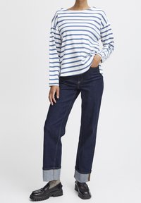 Gestreiftes langärmliges Oberteil in Weiß und Blau, kombiniert mit dunklen Jeans. Die Schuhe sind schwarz, mit dicker Sohle, glänzendem Finish und umgeschlagenem Detail.
