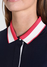 Maglietta blu navy con colletto a coste, presentante strisce rosa e bianche, con bottoni al collo e una patta frontale con cuciture bianche.