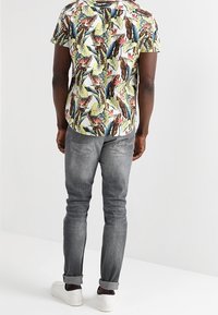 Camicetta a maniche corte con stampa floreale verde, rossa e gialla su sfondo bianco, abbinata a jeans grigi slim-fit e sneakers bianche.