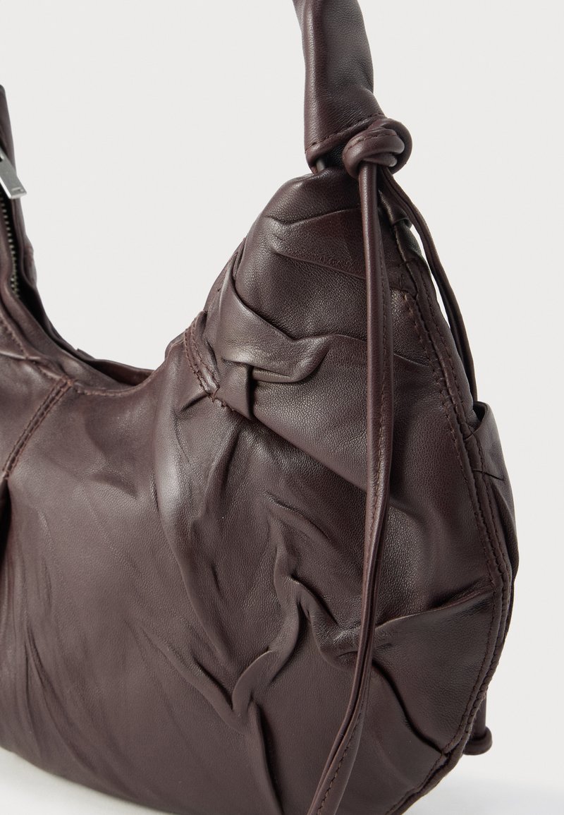 Brun læderhåndtaske med en tekstureret, rynket finish. Har en enkelt knudet strop og en lynlåslukning. Rundet, asymmetrisk form.