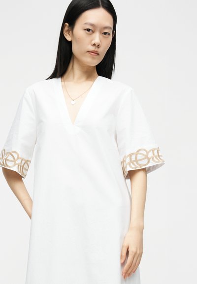 Femme aux longs cheveux noirs portant une robe blanche à col en V avec des boucles brodées beiges sur les manches courtes, sur un fond uni.