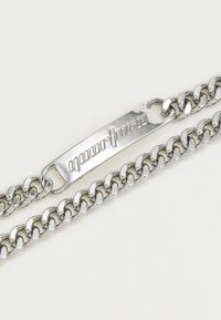 Silverfärgad metallkedjearmband med en platt, graverad platta. Har sammankopplade länkar och en glänsande finish.