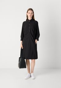 Zwarte jurk met een hoge kraag, halve rits aan de voorkant, gathered zijdetails en zakken. Gepaarde met witte sneakers en een zwarte tas.