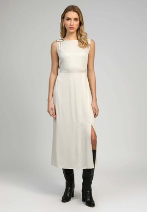 Femme debout portant une robe midi crème sans manches avec une fente latérale et des bottes noires montantes jusqu'aux genoux, sur un fond uni.