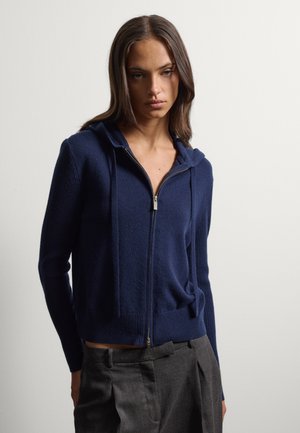 Jonge vrouw die een donkerblauwe hoodie met rits en losse grijze broek draagt, staand tegen een effen lichte achtergrond.