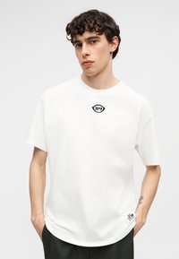 Jeune homme aux cheveux bouclés portant un T-shirt blanc avec un petit logo "R9" sur la poitrine et un patch rectangulaire près de l'ourlet, les mains dans les poches.