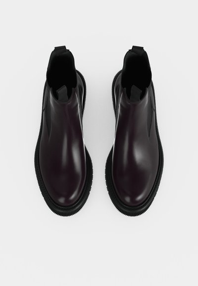 Paire de bottines Chelsea en cuir marron foncé brillant avec semelles épaisses noires rainurées sur fond blanc.