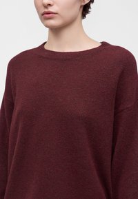 Personne portant un pull tricoté rouge foncé avec un col rond, visible des épaules au menton sur un fond uni.