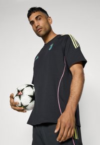 T-shirt de sport noir avec des accents turquoise et jaunes, manches courtes et piping rose, tenu avec un ballon de soccer noir et blanc.