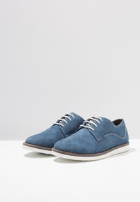 Scarpe da uomo in suede blu con lacci bianchi e cuciture a contrasto, caratterizzate da un design elegante e una suola piatta in gomma bianca.