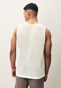 Top smanicato beige chiaro con motivo a rete a diamante, caratterizzato da una scollatura rotonda e una vestibilità comoda, abbinato a pantaloni marroni.