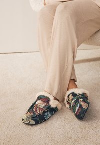 Hausschuhe mit Blumenmuster in Dunkelblau und Kunstfellbesatz. Weicher, strukturierter Stoff mit spitz zulaufendem Design. Gelbe, pinke und grüne Akzente.