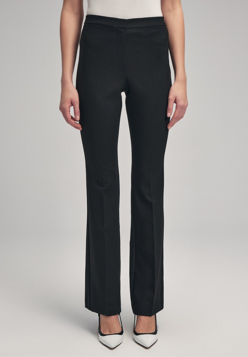 Pantalon noir ajusté avec un design droit, texture lisse, taille mi-haute et détails de couture à l'avant, associé à des chaussures blanches à bout pointu.