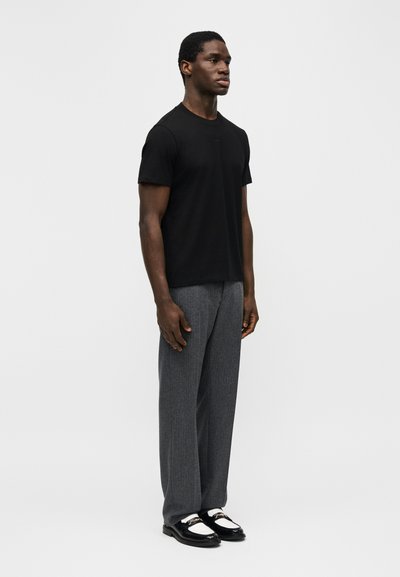 Άνδρας που στέκεται φορώντας μαύρο πουκάμισο με κοντά μανίκια, γκρι παντελόνι και μαύρα λευκά μοκασίνια, μπροστά σε απλό ανοιχτό φόντο.