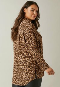 Blusa de leopardo con mangas largas, esquema de colores marrón y beige, textura sedosa, aberturas laterales y dobladillo redondeado.
