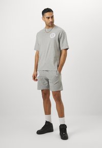 Camiseta de algodón gris y pantalones cortos a juego con un logo en el pecho y la espalda. Se lleva con zapatillas altas negras y calcetines blancos.