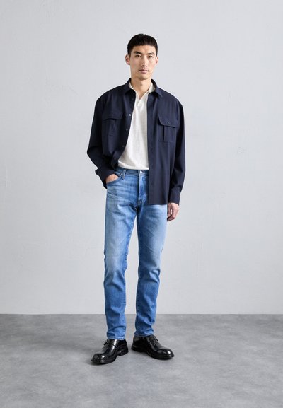 Námornícky dlhý rukáv košele s gombíkmi, béžová polo košeľa, svetlo modré džínsy a čierne formálne topánky. Model stojí na šedom pozadí.