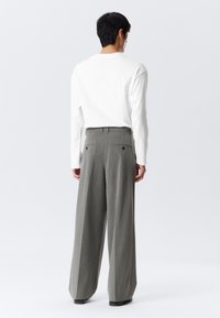 Homme portant un pantalon gris à rayures larges et une chemise blanche à manches longues, se tenant de dos contre un fond blanc uni.