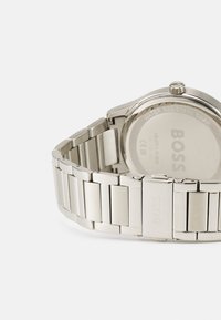 Reloj de acero inoxidable con pulsera plateada pulida y caja redonda, que presenta el nombre de la marca grabado y detalles de resistencia al agua en la parte posterior.