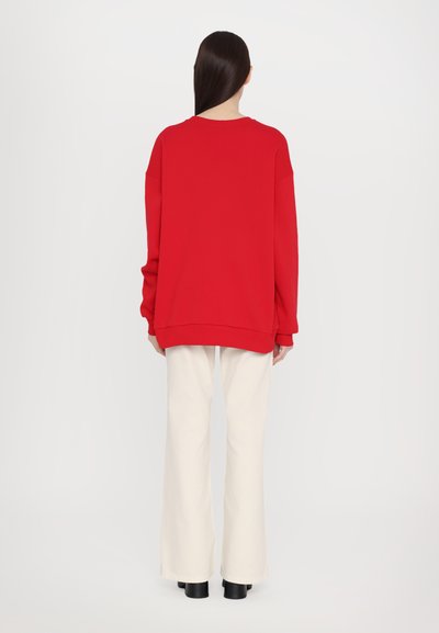 Sudadera roja de gran tamaño con hombros caídos, puños acanalados y un escote redondeado, combinada con pantalones blancos de pierna ancha y zapatos negros con tacón cuadrado.