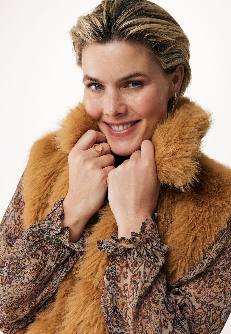 Collar de piel sintética en un rico naranja dorado, enmarcando el rostro. La modelo lleva una blusa estampada con puños con volantes, mostrando detalles suaves y texturizados.