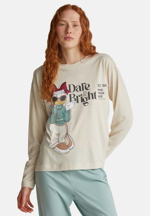 Una giovane donna con lunghi capelli rossi indossa una maglietta a maniche lunghe beige con una grafica di Daisy Duck e il testo "Dare to Be Bright", abbinata a pantaloni azzurri chiari.