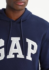 Sweat à capuche bleu marine avec logo "GAP" gris, en coton, capuche avec cordon de serrage et poche kangourou. Texture douce et design décontracté.