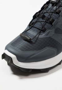 Grijze sportieve schoen met een bovenwerk van textuurstof, elastisch vetersysteem, rubberen buitenzool met gripnoppen en een witte tussenzool.