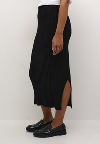 Jupe noire en tricot côtelé avec taille haute et fentes latérales, associée à des mocassins en cuir noir laqué, présentant une texture lisse et une coupe ajustée.