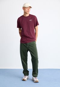 Homem a usar um chapéu bege, uma t-shirt burgundy da Nike, calças de cerzido verdes e ténis brancos, em pé sobre um chão azul contra uma parede branca.