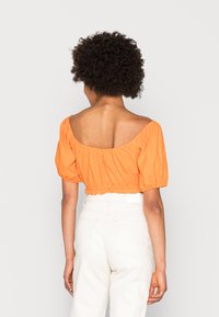 Blusa curta laranja com design off-shoulder e mangas bufantes. Tecido suave, bainha elástica e detalhes franzidos elegantes. Combinada com calças brancas.