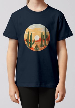 Kind trägt ein marineblaues T-Shirt mit einer kreisförmigen Wüstenlandschaft, die hohe grüne Kakteen, orangefarbene Felsen und eine untergehende Sonne zeigt.
