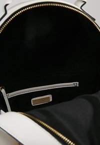 Intérieur noir d'un sac à main blanc avec une fermeture éclair dorée, comprenant une poche zippée et une petite étiquette de marque à l'intérieur.