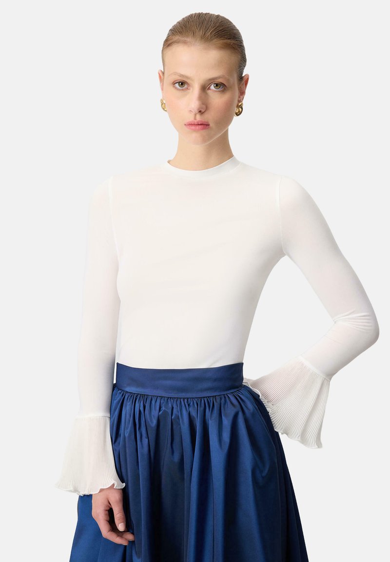 Valkoinen pitkähihainen paita, jossa on pleated, liehuvat hihansuut, yhdistettynä siniseen, korkeavyötäröiseen hameeseen, jossa on poimutettu vyötärö. Pintakäsittely on yleisesti sileä.