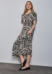 Zwart en beige gestreepte jurk met korte mouwen, een ingezaagde taille en een vloeiende rok. Gecombineerd met zwarte flats, tegen een neutrale achtergrond.