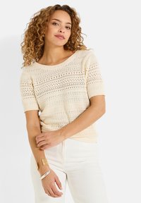 Top de punto de color crema, de manga corta, con un patrón texturizado de trabajo abierto. Presenta un escote ancho y bordes acanalados.