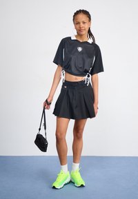 Svart kortärmad tröja med nätstruktur och vit krage; svart veckad kjol; neongröna sneakers; liten svart handbag.