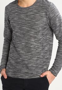 Man med lång ärm, svart och vit melerad stickad tröja, med händerna i fickorna på mörka byxor, stående mot en enkel bakgrund.