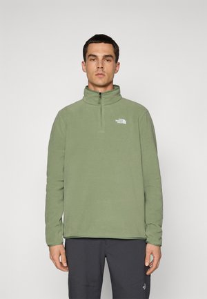 Mann iført olivenfarget fleecegenser med kvart glidelås og The North Face-logo på brystet, samt svarte bukser, mot en enkel lys bakgrunn.