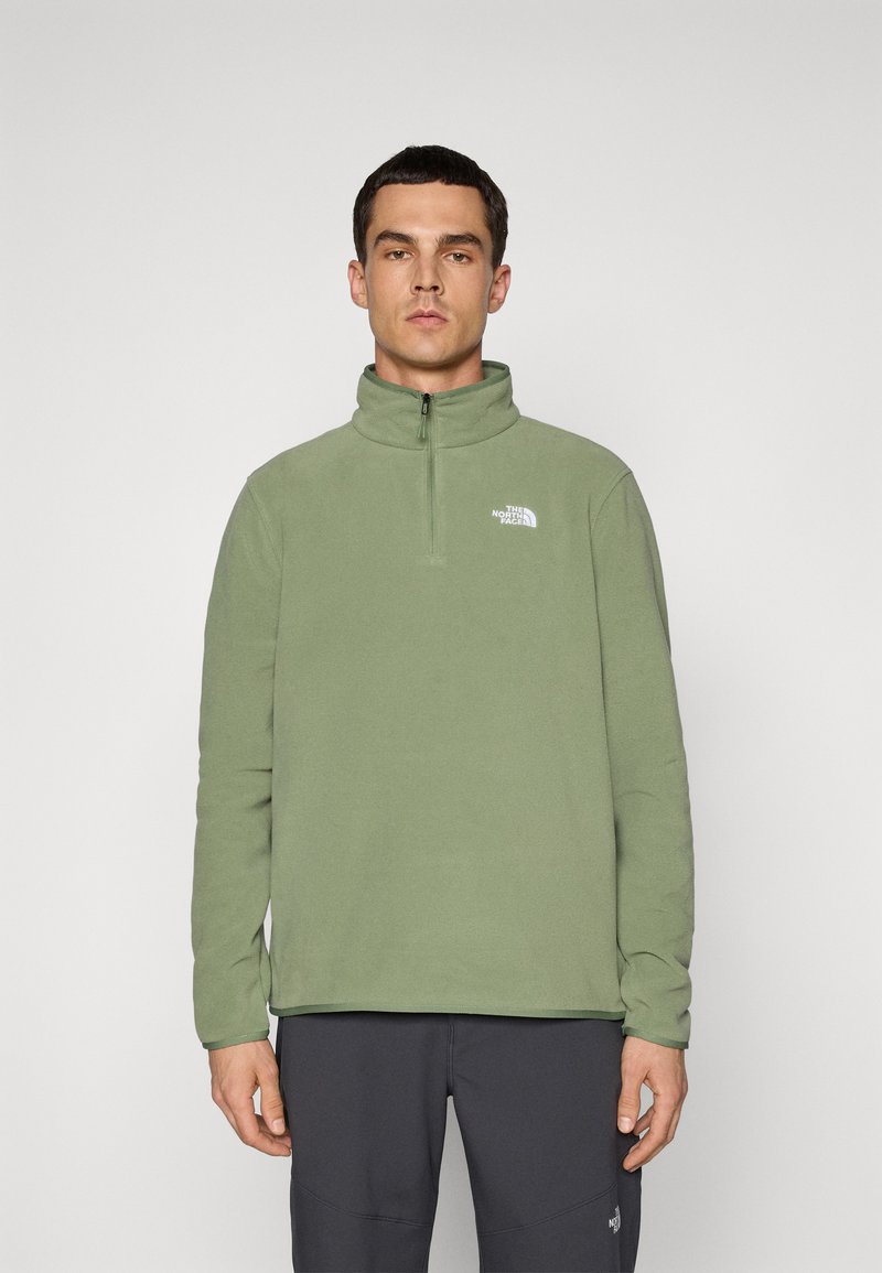 Muškarac u maslinasto zelenoj polar jakni s četvrtim patentnim zatvaračem i logom The North Face na prsima, u crnim hlačama, na jednostavnoj svijetloj pozadini.