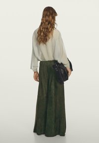 Zwiewna, jasnozielona bluzka z bufiastymi rękawami, w połączeniu z długą, ciemnozieloną spódnicą z zamszu. Czarna torebka na ramię spoczywa w dłoni modelki.