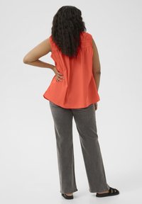 Top sans manches orange avec des épaules froncées, associé à un pantalon large gris et des sandales noires. Le tissu semble léger et lisse.