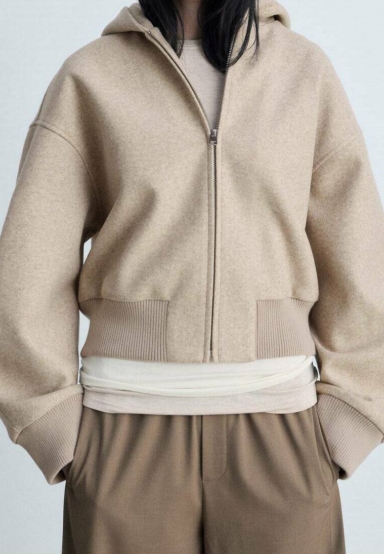 Sudadera con cremallera en color beige, fabricada en un tejido suave, con dobladillo y puños acanalados. Lleva puesta sobre una camiseta clara, combinada con pantalones marrones.