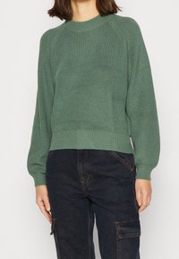 Maglione verde lavorato a maglia con una texture a coste, scollatura rotonda e maniche raglan. Abbinato a pantaloni cargo scuri, che mostrano un fit casual.