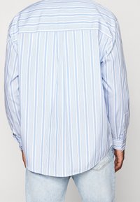 Světle modrá pruhovaná košile s dlouhými rukávy, obsahující svislé pruhy v různých odstínech. Měkká tkanina s zaobleným lemem a manžetami na knoflíky.