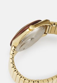Montre en métal doré avec une lunette marron, couronne texturée et bracelet doré à maillons. Le dos est en acier inoxydable avec une finition lisse.