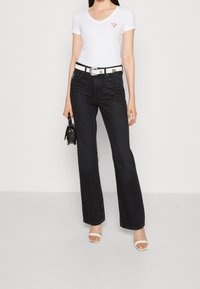 Camiseta blanca de cuello en V con un logo rojo, combinada con jeans negros de cintura alta y acampanados, y un cinturón blanco, complementada con sandalias de tacón claro tipo bloque.