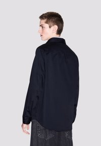 Camicia blu navy con maniche lunghe, dotata di colletto standard e texture liscia. La vista posteriore mostra le cuciture delle spalle e l'orlo curvo.